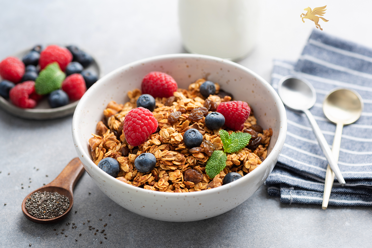 Acovior Low Sugar Simply Granola served in a white bowl with fresh raspberries, blueberries, and mint leaves, alongside chia seeds and milk on a light grey background.

